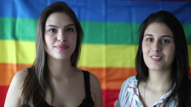 Two young women posing to camera with LGBT rainbow flag in background
