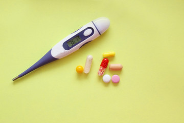 Multi-colored tablet and electronic thermometer lie on yellow background in the center. Drug use. Treatment. Home first aid kit. Disease. Emergency services. View from above. Pain. opy space for text