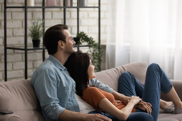 Smiling millennial Caucasian couple sit rest on couch at home hug and cuddle look in distance dream of happy future together, young man and woman tenants relax on comfortable sofa in living room