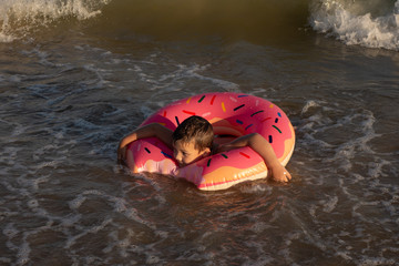 A 5-year-old boy is swimming and having fun in the sea near the shore with a donut-shaped inflatable circle.