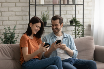 Obraz premium Happy young Caucasian couple sit relax on sofa at home use smartphones gadgets together, smiling millennial man and woman rest on couch in living room have fun browsing cellphone, technology concept