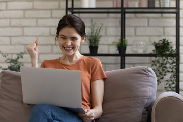 Overjoyed millennial girl sit relax on couch at home feel euphoric read good news on computer,...