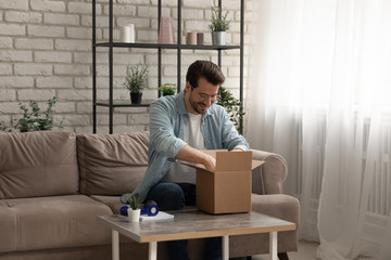 Happy millennial caucasian man sit on sofa at home open unbox carton package with internet order,...