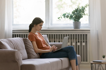 Focused young Caucasian woman sit on sofa in living room work on laptop check write email on gadget, millennial female look at computer screen, browse wireless internet on device, technology concept