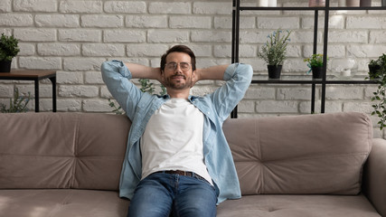 Happy young Caucasian man sit relax on comfortable sofa at home breathe fresh air relieve negative...