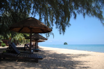 東南アジアのマレーシアにあるトロピカルなリゾートのティオマン島の木陰のビーチにあるパラソルとサマーベッドと向こうに見える青い海と小島