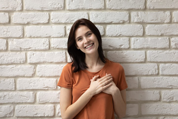 Fototapeta na wymiar Portrait of smiling young Caucasian woman isolated on white brick wall background hold hands at heart chest show gratitude support, happy millennial girl volunteer feel thankful demonstrate care