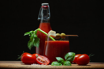 Fresh tomato juice in a glass and bottle with branch of fresh celery,basil leaves,green olives,piece of parmesan cheese and tomatoes on wooden table.Close up of healthy drink.