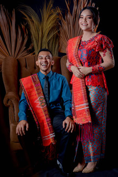 portrait of beautiful indonesian couple wearing traditional batak costume sit on brown couch isolated on black background.