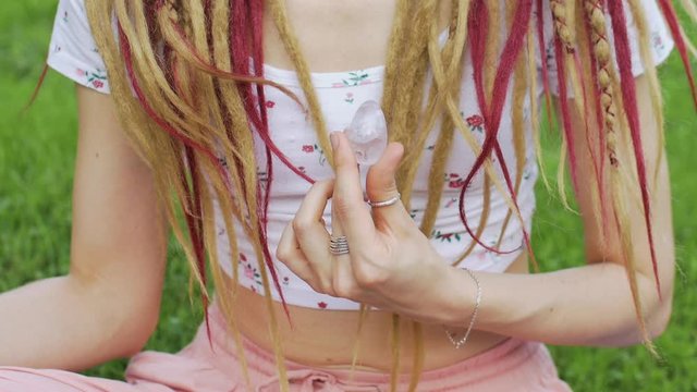 Adult girl with long dreadlocks is holding transparent violet amethyst yoni egg for vumfit, imbuilding or meditation outdoors on her body background outdoors