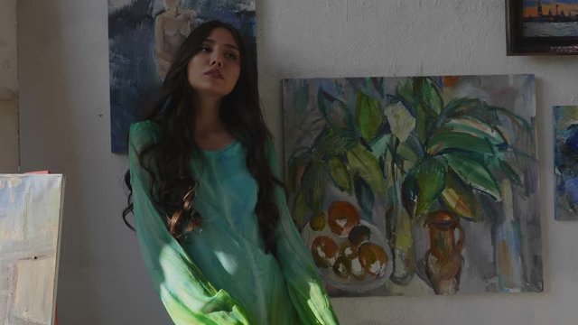 attractive girl with long curly hair wearing green dress poses near own pictures on wall in art studio slow motion