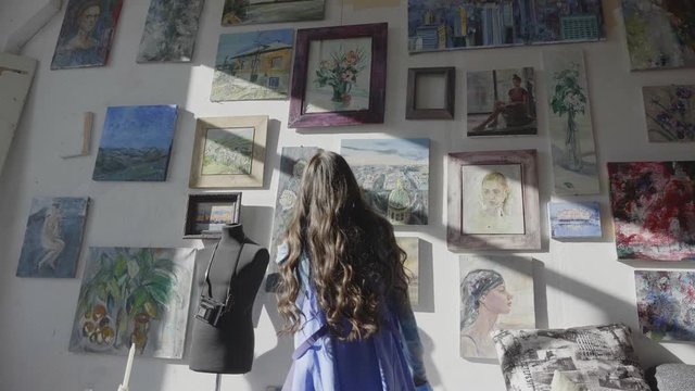 curly haired lady ballerina in long blue dress dances near wall with paintings in art studio backside view slow motion