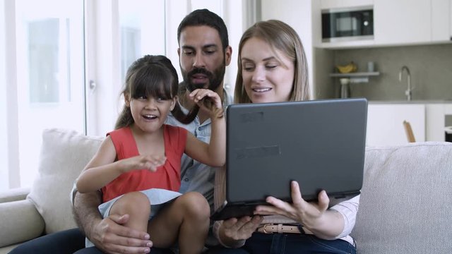 Cheerful adorable girl sitting on fathers lap, waving hello, showing hair and speaking at laptop webcam. Happy family using computer for video call. Medium shot. Communication concept