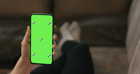 young woman lying on a couch and holding smartphone with vertical green screen
