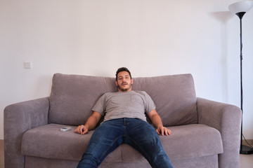 Portrait of a bored and tired young man sitting on a sofa watching television absorbed with his...