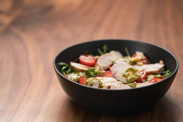 Salad with chicken, tomatoes and arugula in black bowl on walnut wood background with copy space