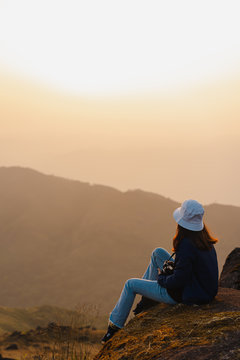 A woman with a red long-sleeved shirt and a white hat sits on the large rocks at Mulayit Taung, Myanmar. She holds a camera and looks forward to the overlapping hillsides and golden dry grasslands.