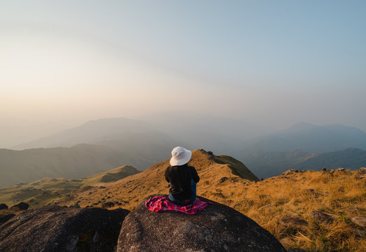 A woman with red long-sleeved shirt and a white hat sits back on a large canyon at the high peak of Mulayit Taung, Myanmar. There are overlapping hills and golden dry grasslands. There is copy space.