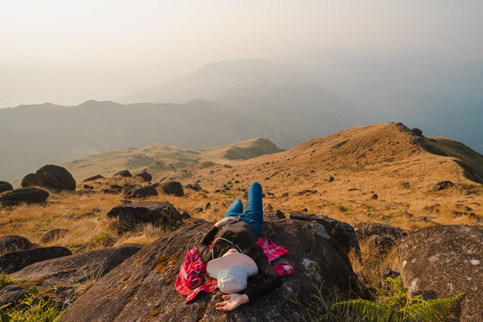 A woman with black long-sleeved shirt and a white hat lie down on a large stone at Mulayit Taung, Myanmar. There are overlapping hills and golden dry grasslands with a warm tone. There is copy space.