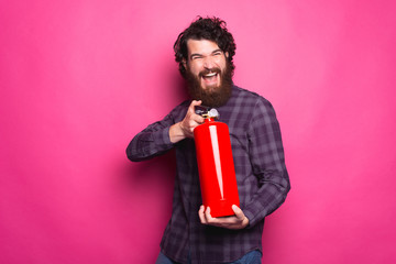 Photo of young bearded man in shirt holding fire extinguisher over pink background.
