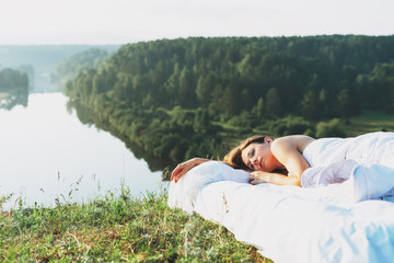 Young woman lying in bed with white bed linen in nature against beautiful landscape early morning
