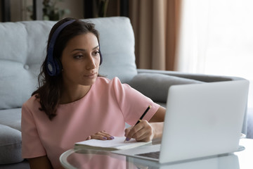 Concentrated young Arabic indian woman in headphones look at laptop screen make notes watching...