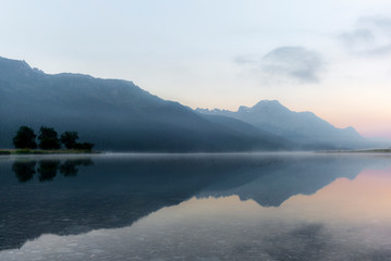 The mountains reflecting in the lake of Silvaplana in the Engadin valley at sunrise with the fog over the water