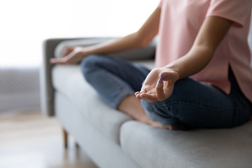 Crop close up of woman sit on couch at home practice yoga in lotus position with mudra hands, calm relaxed female rest on sofa indoors, meditate relieve negative emotions, stress free, peace concept