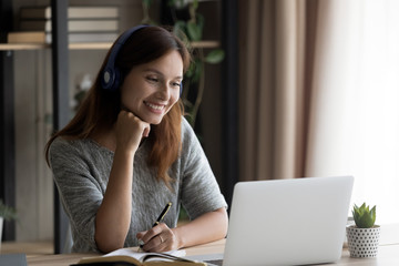 Happy young red-haired Caucasian woman in headphones sit at desk at home look at laptop screen watch webinar, smiling female in earphones study online on computer, take distant education course