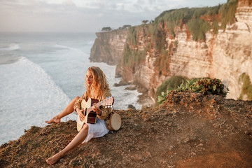 Happy girl with straw hat is sitting on sea cliff with guitar, enjoy nice view on the ocean and play guitar