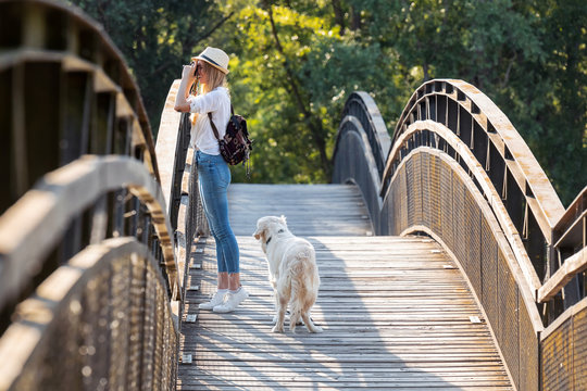Young amateur photograph woman taking a photo of the landscape while walking with her dog crossing over a bridge in the park.