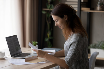 Smiling young Caucasian woman sit at table at home open envelope read paper correspondence notice,...