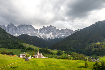 Wide angle view of the Val Di Funes in northern Italy, with a small church surrounded by meadows and trees in the foreground and a mountain range in the background, under a dark sky with stormy clouds