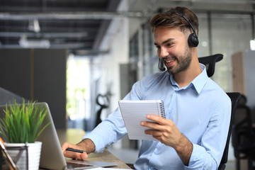 Fototapeta na wymiar Smiling male business consultant with headphones sitting at modern office, video call looking at laptop screen. Man customer service support agent helpline talking online chat.