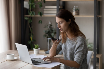 Focused young Caucasian woman sit at desk at home work on laptop record audio message on cellphone, millennial female use computer browsing, activate virtual digital voice assistant on smartphone