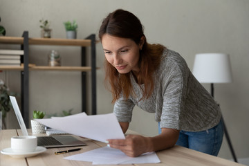 Focused young Caucasian businesswoman lean at table at home consider paper documents work on computer, millennial woman busy using laptop analyzing paperwork correspondence at workplace
