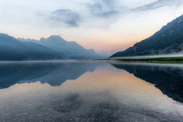 A fisherman rowing among the fog on the lake of Silvaplana in the Engadin valley at sunrise with mountains reflecting in the water