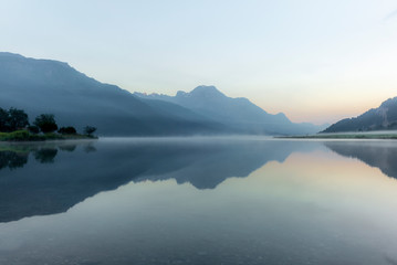 The mountains reflecting in the lake of Silvaplana in the Engadin valley at sunrise with the fog over the water