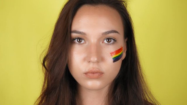 A woman with a rainbow pattern on her face is looking at the camera. The LGBT flag is painted on the face. Yellow background.