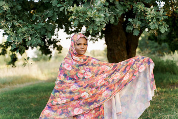 Muslim black woman African ethnicity weared traditional colorful hijab smiles under green tree. Arabic islam religion clothes. Pretty black girl smiles. Business for black woman.