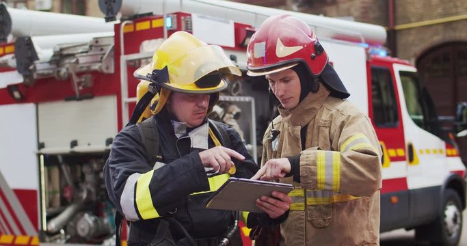 Medium plan of two firefighters discussing rescue plan looking at tablet while standing in helmets near a fire van. The concept of saving lives, heroic profession, fire safety