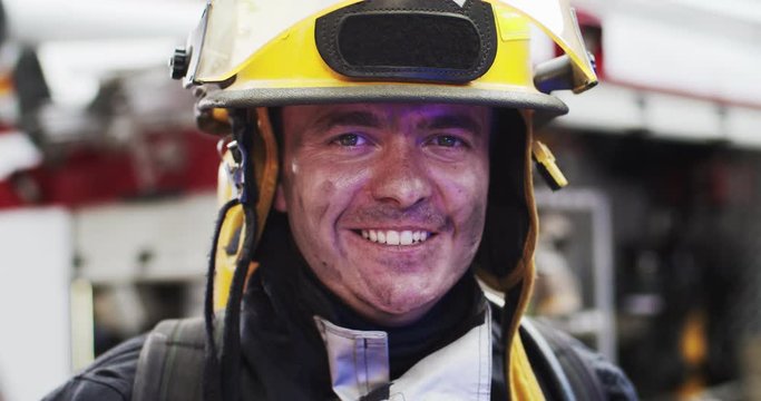 Close up portrait of happy smiling fireman in helmet and gull equipment standing next to car with flashing lights on and looking into camera. Concept of saving lives, heroic profession, fire safety