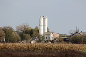 Fototapeta na wymiar Two white old metal storage silos with fence on one side protected with safety net rising above industrial complex surrounded with cornfield and trees on clear blue sky background