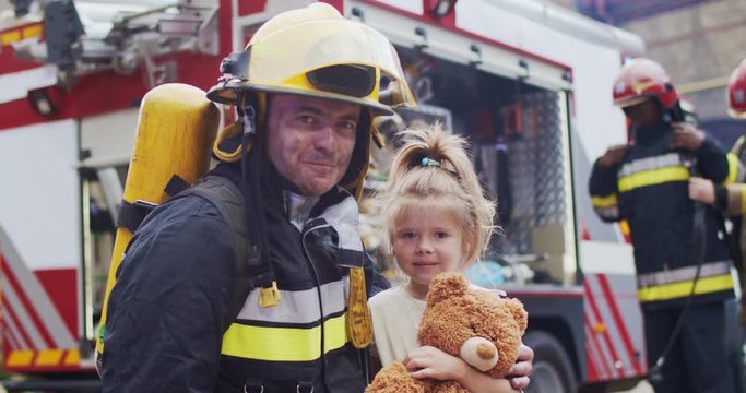 Firefighter hug rescued little girl with teddy bear. Frightened child rejoices in rescue. At background firefighters after extinguishing fire next to fire truck. Concept of saving lives, fire safety