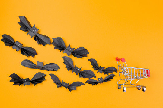 A flock of bats made of paper flies behind a shopping cart. Orange background. Copy space. Flat lay. The concept of Halloween and holiday shopping