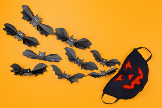 Reusable anti-virus mask with print of jack lantern surrounded by paper bats. Orange background. Copy space. Flat lay. The concept of protection from the virus during Halloween
