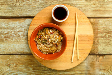 Funchoza with vegetables and mushrooms in a clay plate on a round stand next to chopsticks and soy sauce.