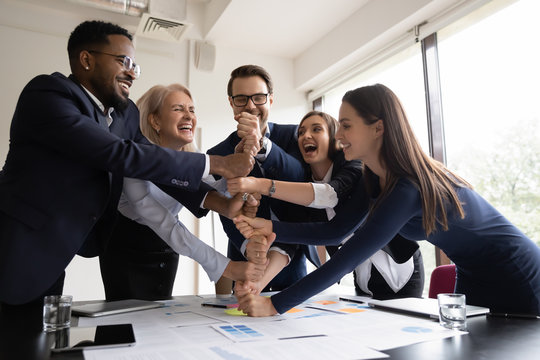 Happy Laughing Businesspeople Colleagues Of Different Age And Race Stacking Fists On Meeting Or Motivation Training In Office Demonstrating Togetherness, Unity, Cooperation And Corporate Team Spirit