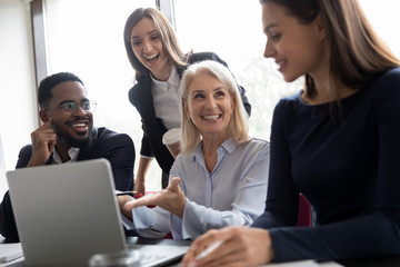 Pleasant mature woman qualified employee explaining business task details to younger colleagues, happy aged lady team leader showing diverse millennial staff solution of problem on computer screen