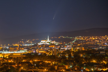 Comet Neowise in the night sky over the illuminated city and mountains in the background